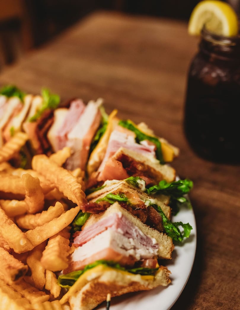 Club sandwiches with fries and a drink with a lemon wedge on a wooden table.