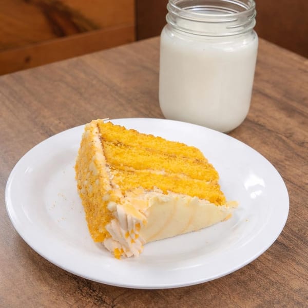 A slice of yellow cake with white frosting and a glass of milk on a wooden table.