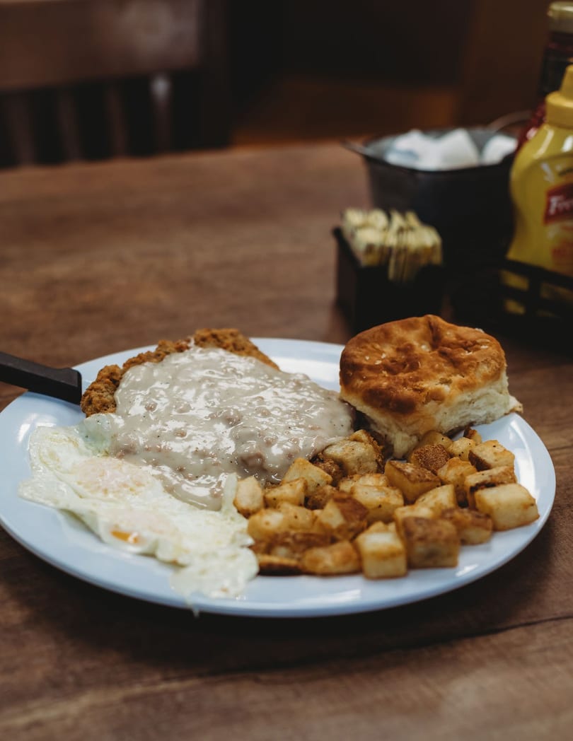 Chicken fried steak with gravy, fried eggs, home fries, and a biscuit on a white plate.
