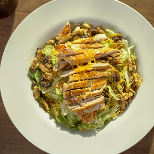 Grilled chicken salad with lettuce, walnuts, raisins, and shredded cheese on a white plate.