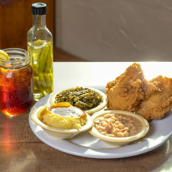 Fried chicken, collard greens, baked beans, grits, iced tea, and pickled peppers on a wooden table.