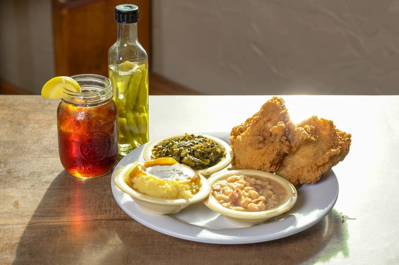 Fried chicken, collard greens, baked beans, grits, iced tea, and pickled peppers on a wooden table.
