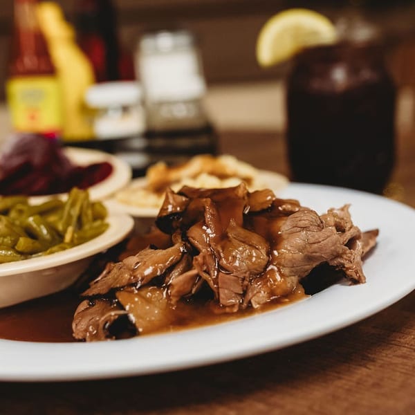 A plate of pot roast with gravy, green beans, and other sides, with a drink in the background.