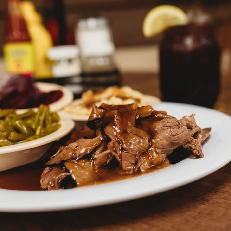 A plate of pot roast with gravy, green beans, and other sides, with a drink in the background.