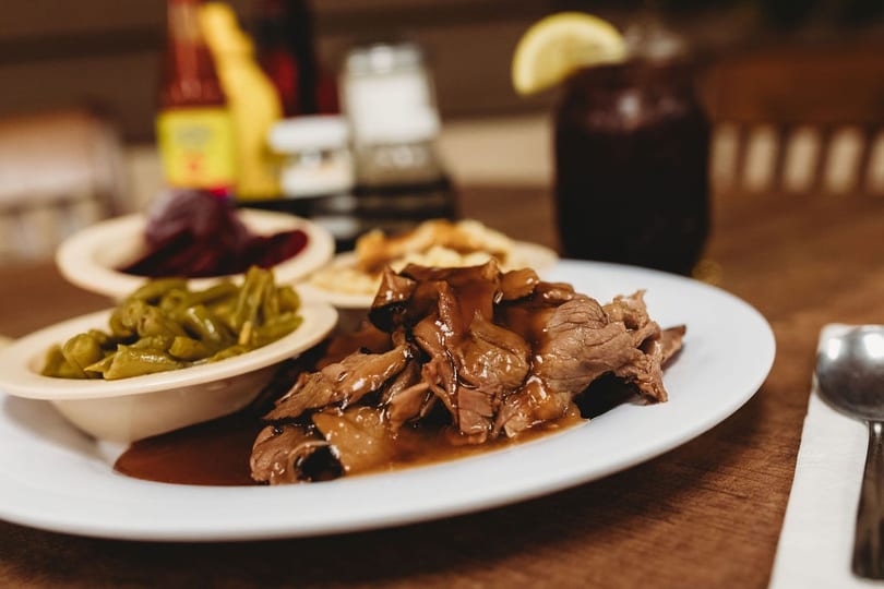 A plate of pot roast with gravy, green beans, and other sides, with a drink in the background.