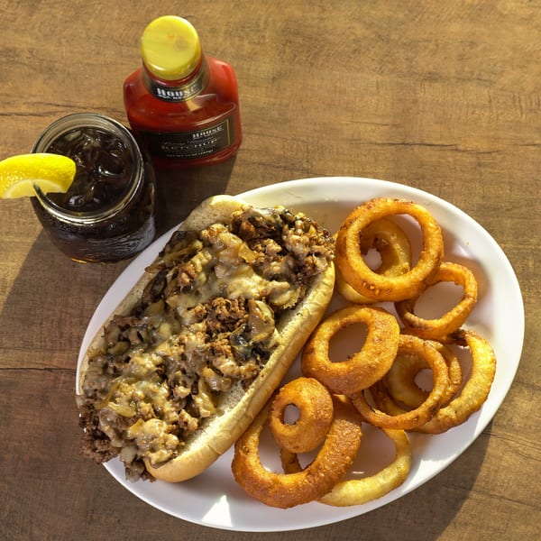 Cheesesteak sandwich with mushrooms and onions, onion rings, soda with lemon, and ketchup on a wooden table.