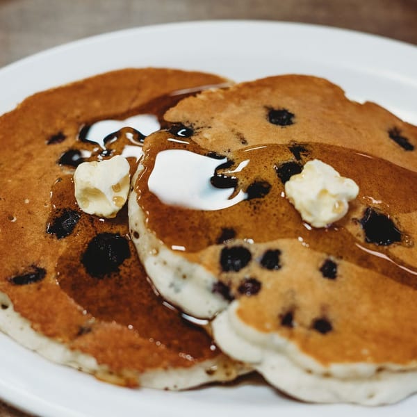 Two blueberry pancakes with pats of butter and maple syrup on a white plate.