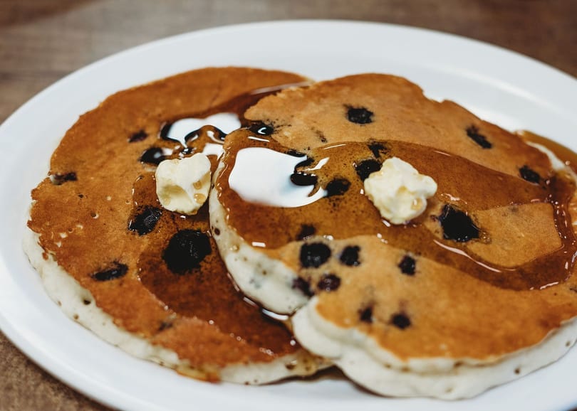 Two blueberry pancakes with pats of butter and maple syrup on a white plate.