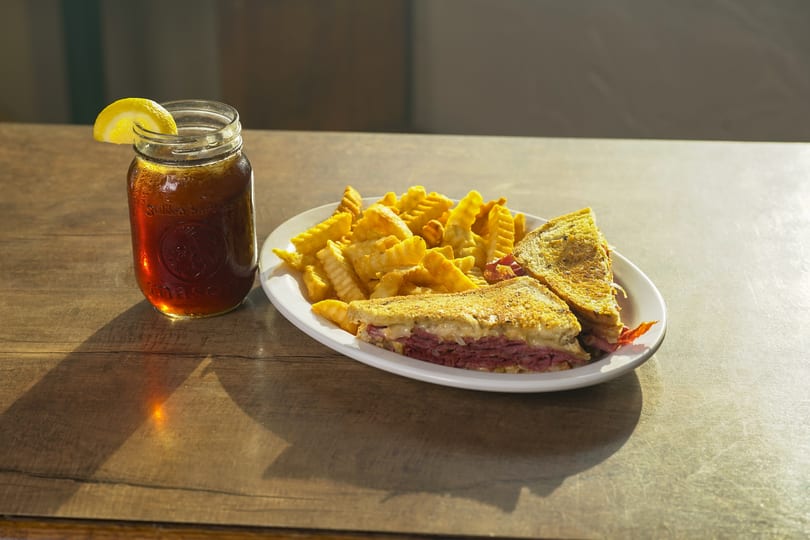 Reuben sandwich with crinkle-cut fries and iced tea with a lemon slice on a wooden table.