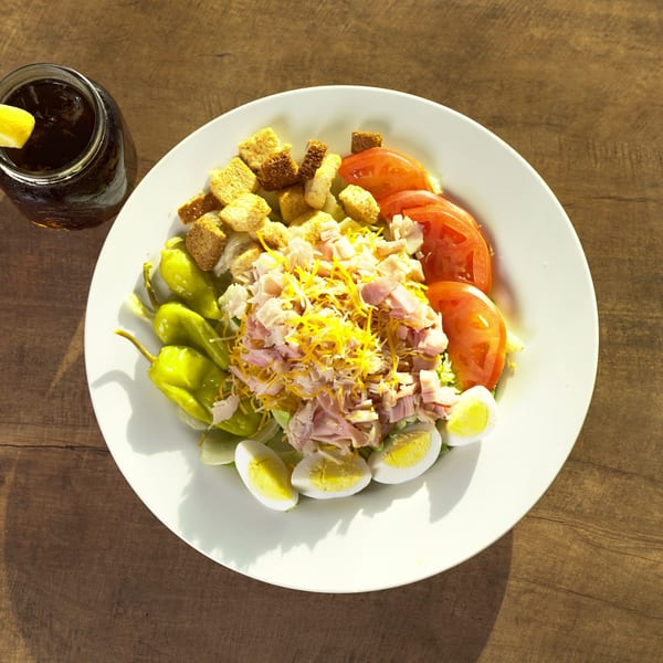 Chef salad with ham, cheese, tomatoes, eggs, croutons, and peppers, with a drink.