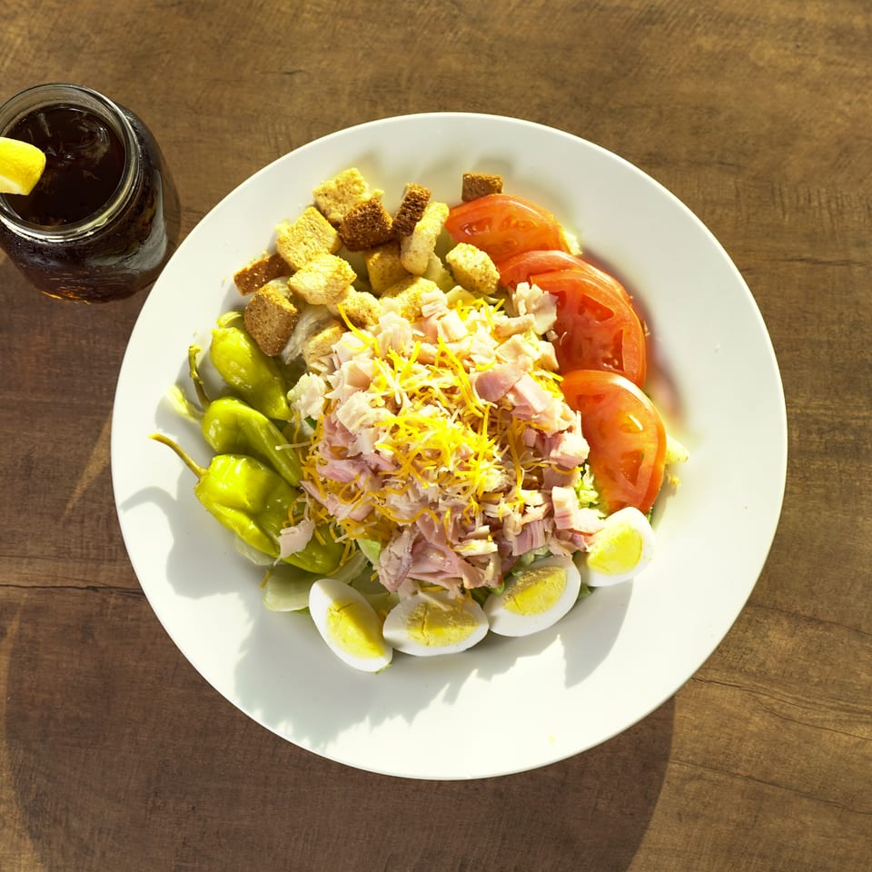 Chef salad with ham, cheese, tomatoes, eggs, croutons, and peppers, with a drink.