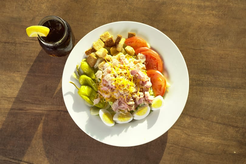 Chef salad with ham, cheese, tomatoes, eggs, croutons, and peppers, with a drink.