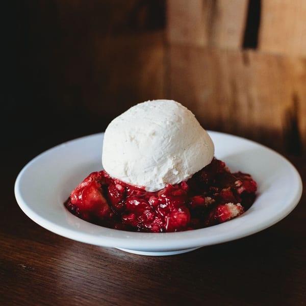 A white bowl holds a scoop of vanilla ice cream atop a vibrant red berry cobbler.