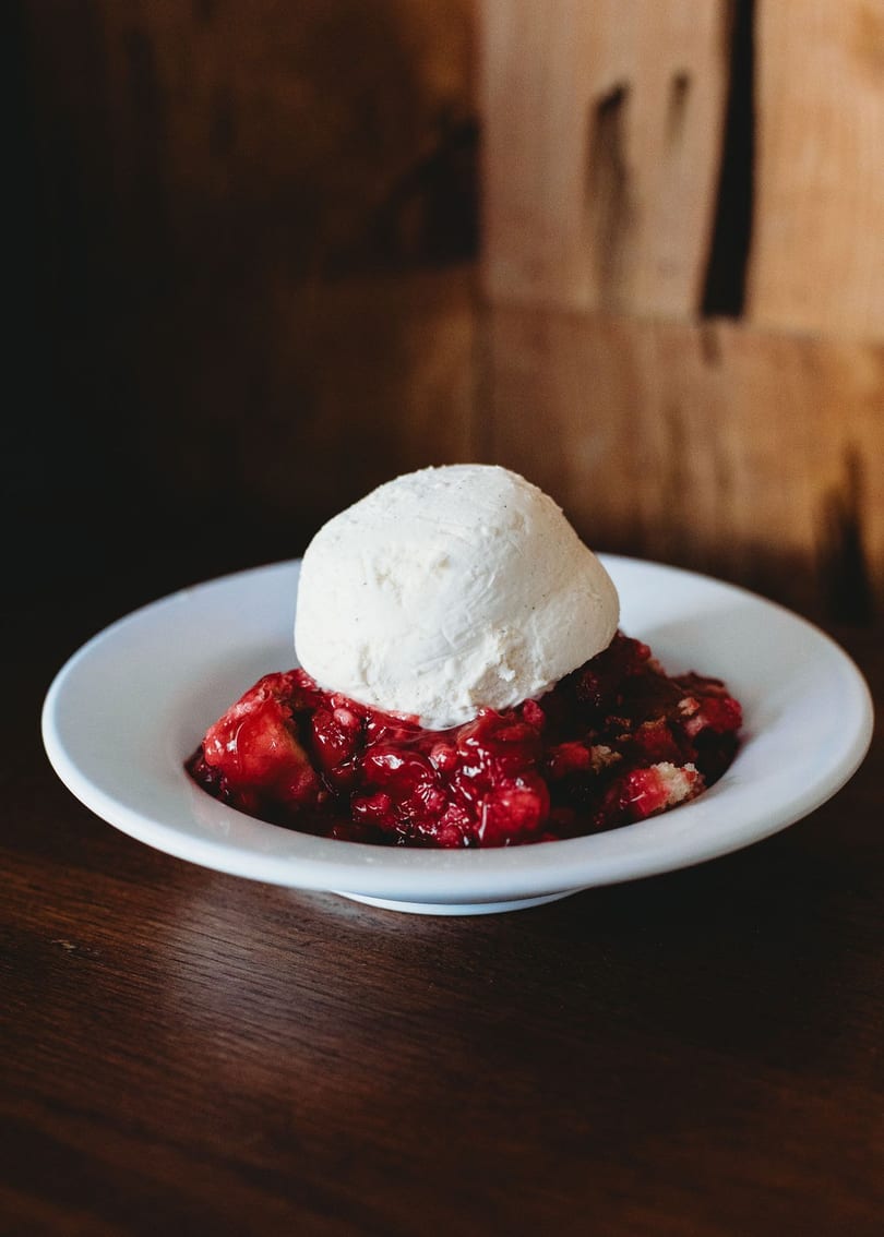 A white bowl holds a scoop of vanilla ice cream atop a vibrant red berry cobbler.